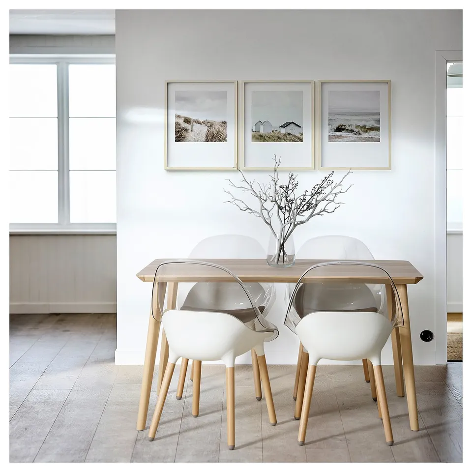 Modern dining space with a light wood table, four white chairs with curved backs, and three framed coastal photos on the wall.