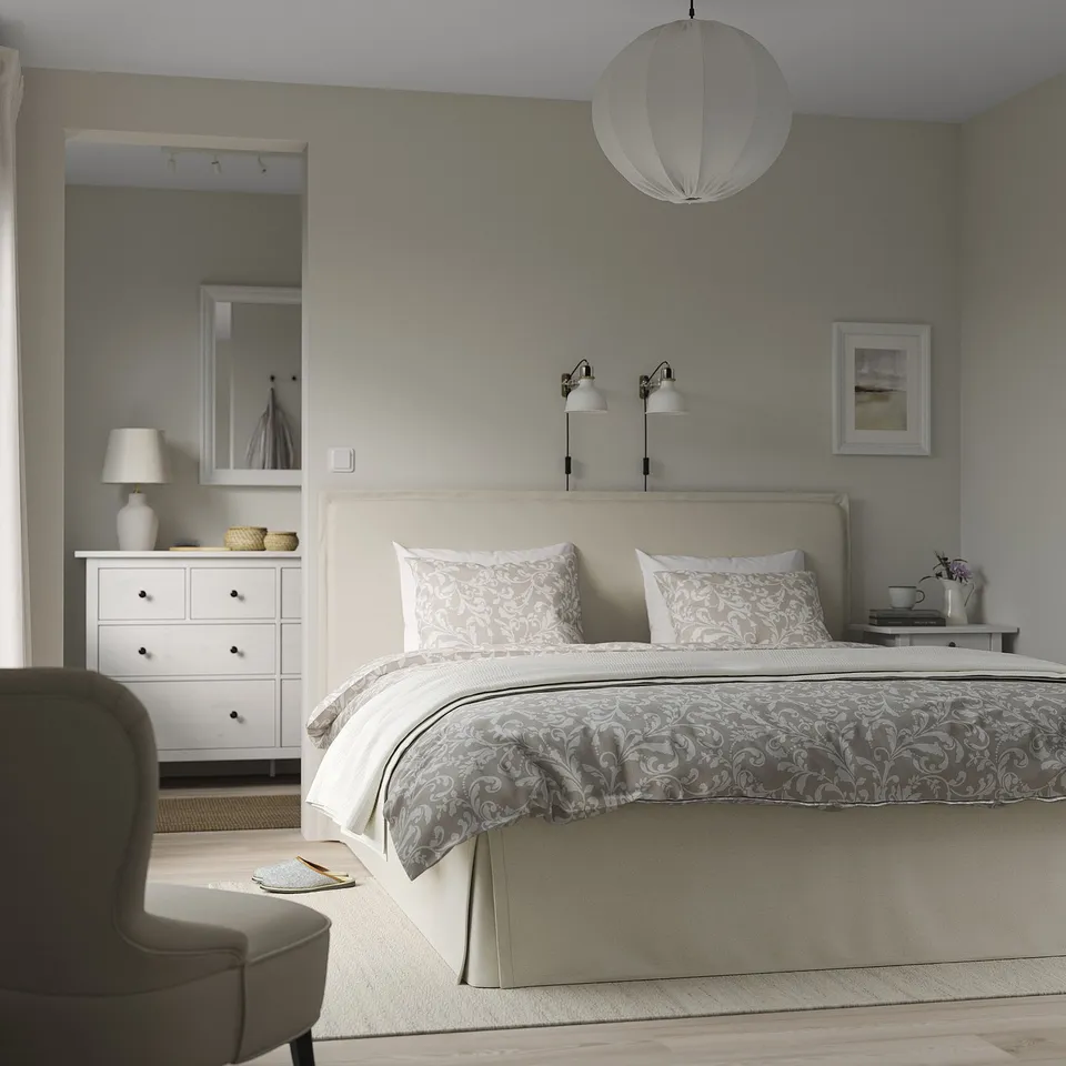 Bedroom with white bed, patterned cover, hanging light, grey chair, white dresser, mirror.