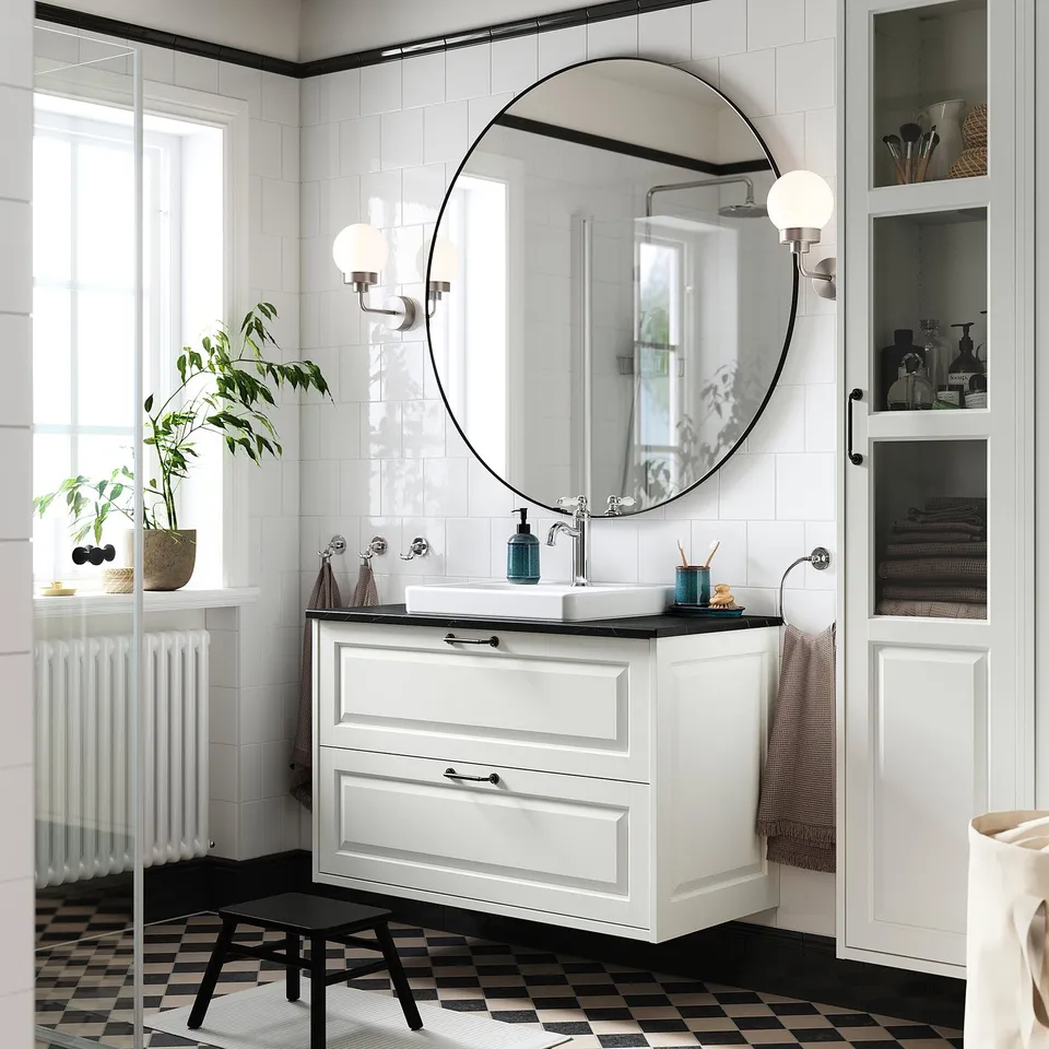 Modern white bathroom. Black tiles, round mirror, TÄNNFORSEN vanity, sink, sconces, tall cabinet, plant.
