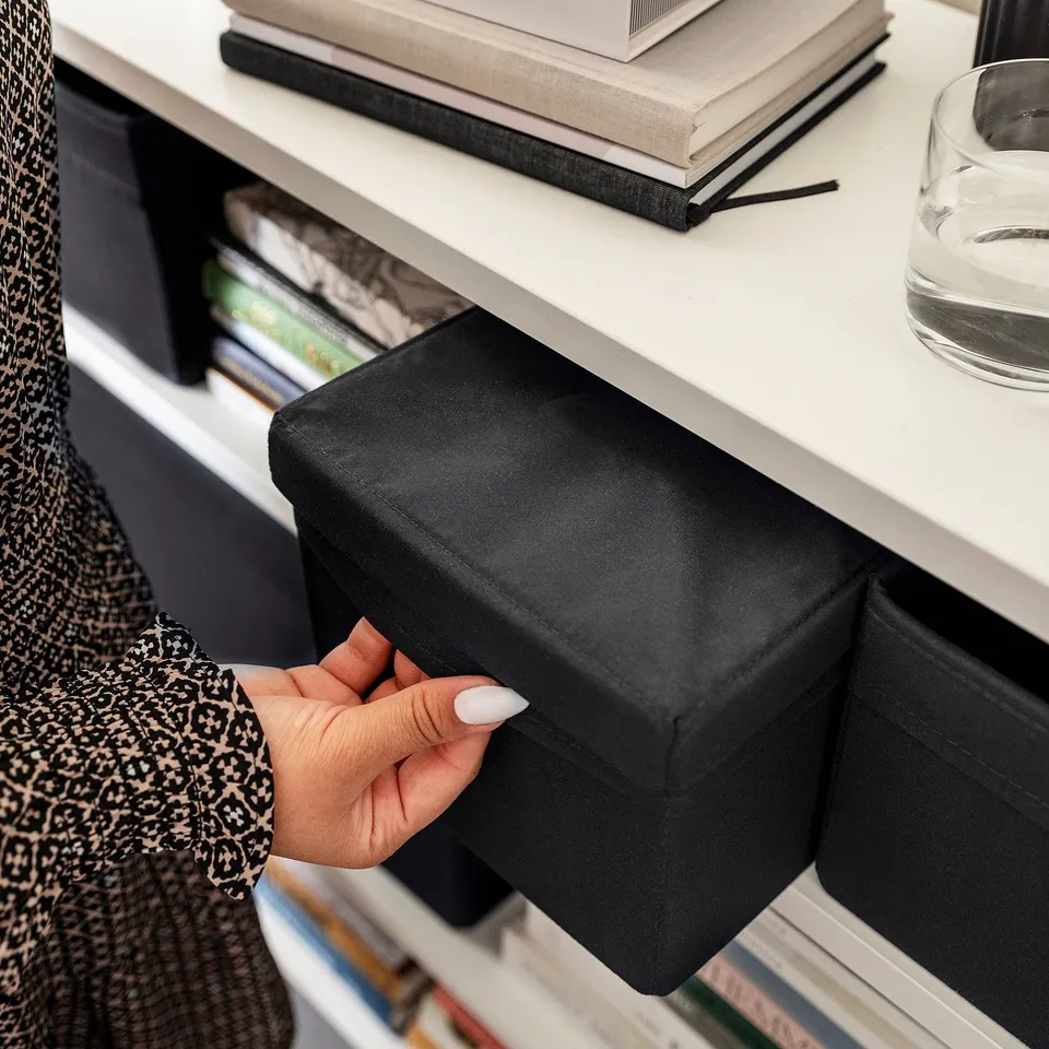 Hand pulls black fabric lid from white shelf, revealing organised storage box.