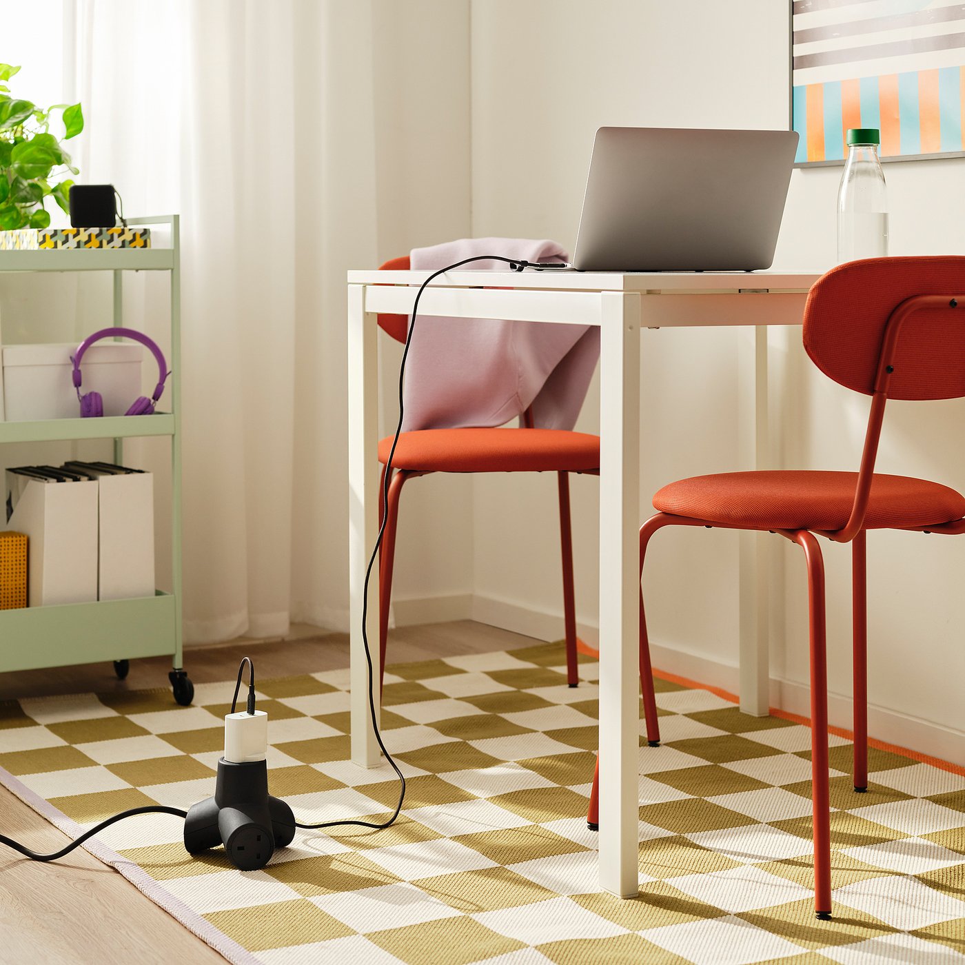 White desk with laptop, chair, purple headphones, green shelving, and a black, 4-way extension cord on the floor. Chequered rug.