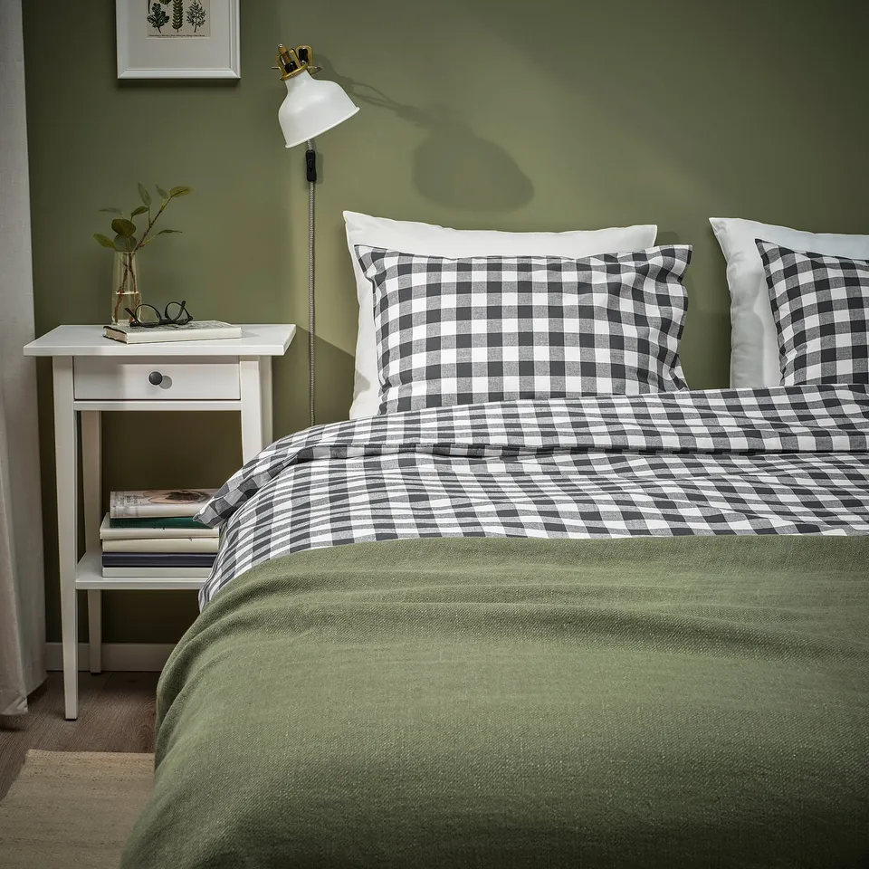 Bed with black & white gingham duvet, green blanket; white bedside table, lamp, vase.