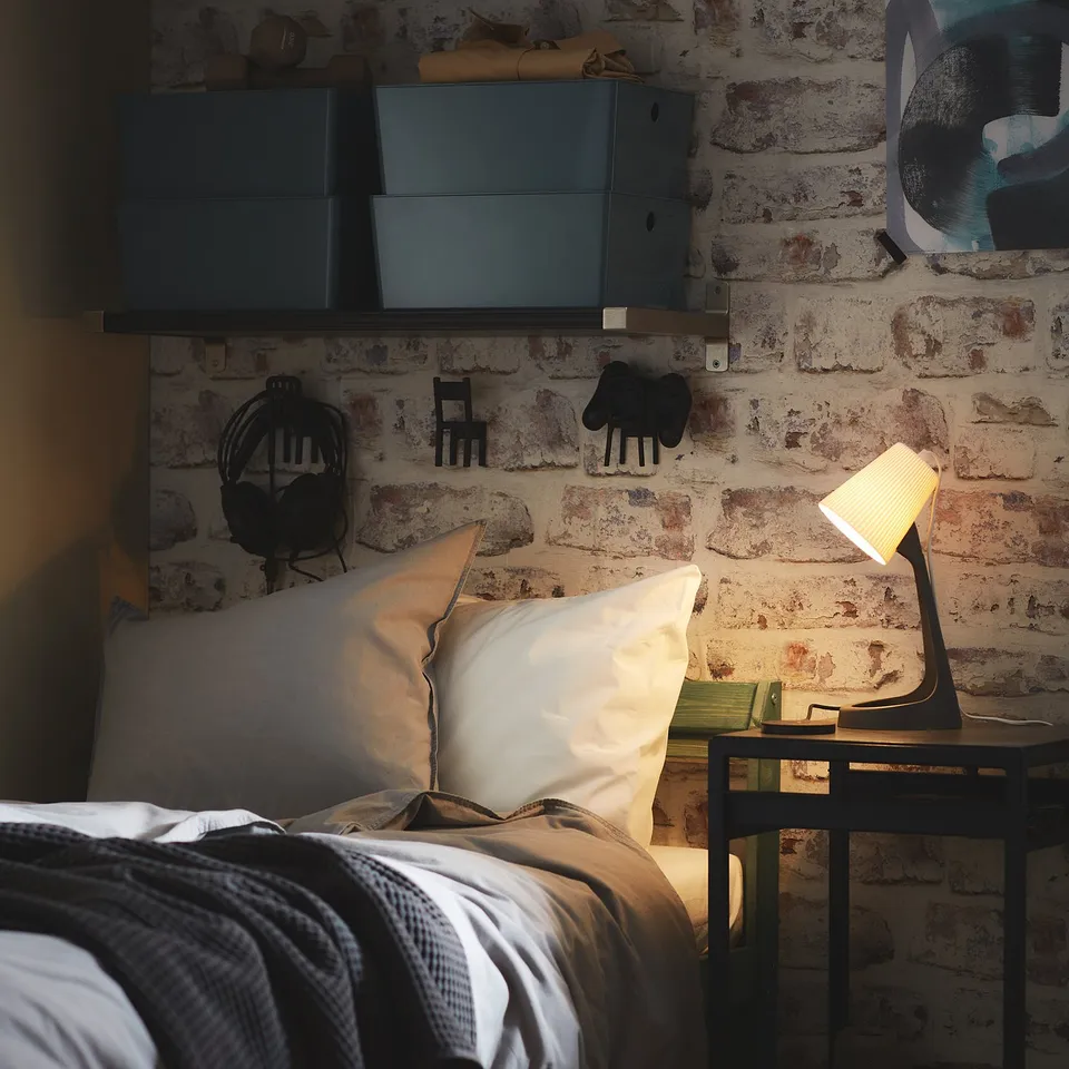 Bedroom with brick wall, white pillows, grey blanket, small black desk, lamp, and wall-mounted cabinets.