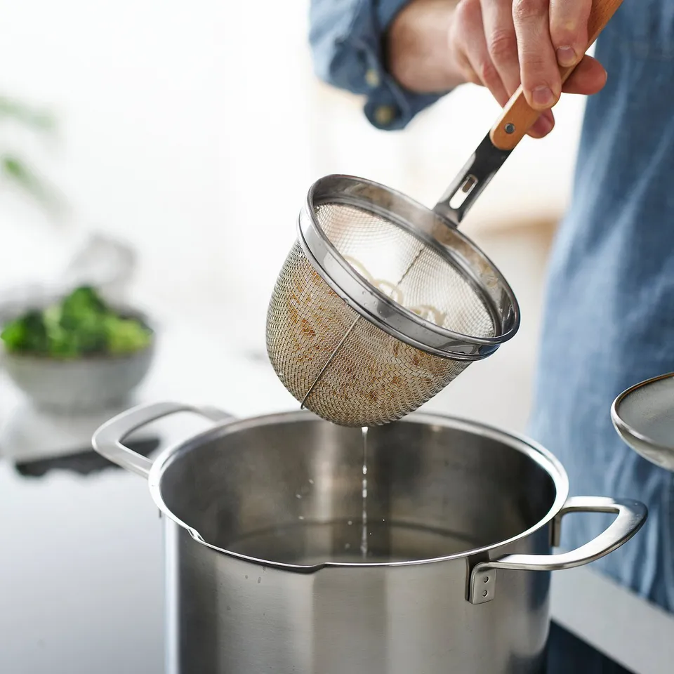 Person straining food in kitchen with VARDAGEN strainer, ensuring noodles stay springy.
