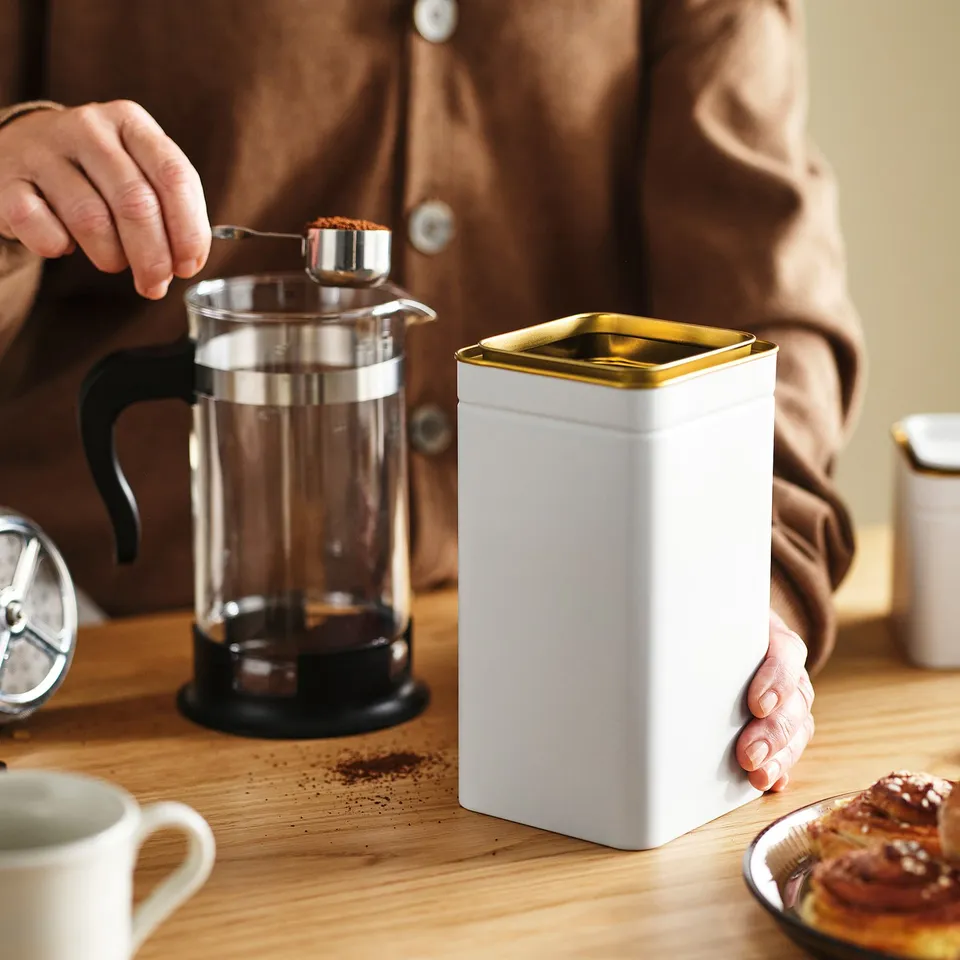 Person scoops coffee grounds into a cafetière from a white metal tin next to a pastry on a wooden table.