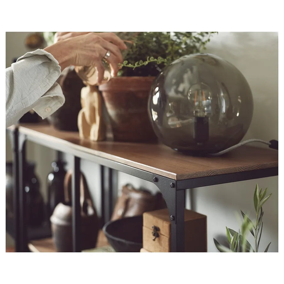 Person decorates shelf with lamp, plants, and wooden figurine. Modern, rustic design with wooden top and metal frame. Green table lamp on right.