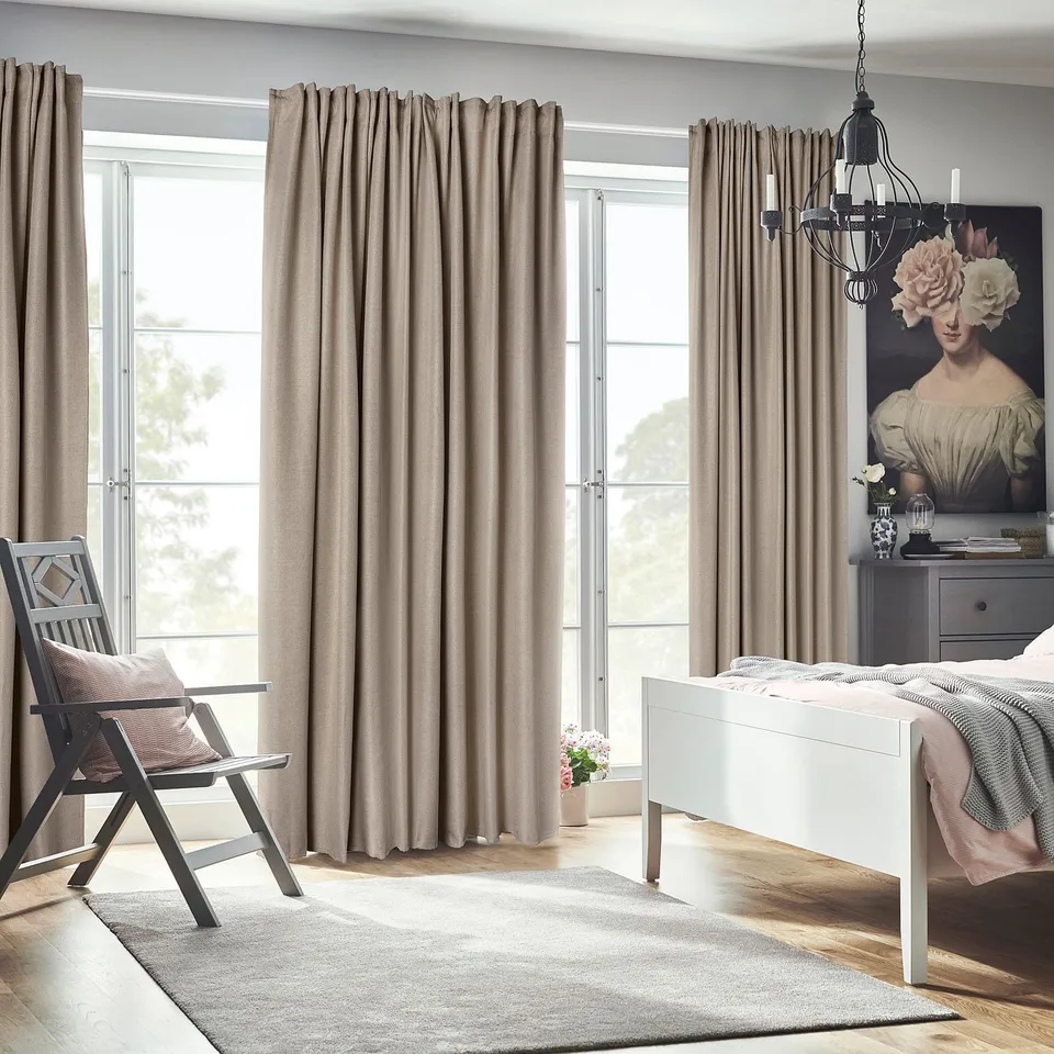 Bedroom with ANNAKAJSA room-darkening beige curtains, a white bed, grey chair, and floral artwork. Curtains reach floor, framing large window.
