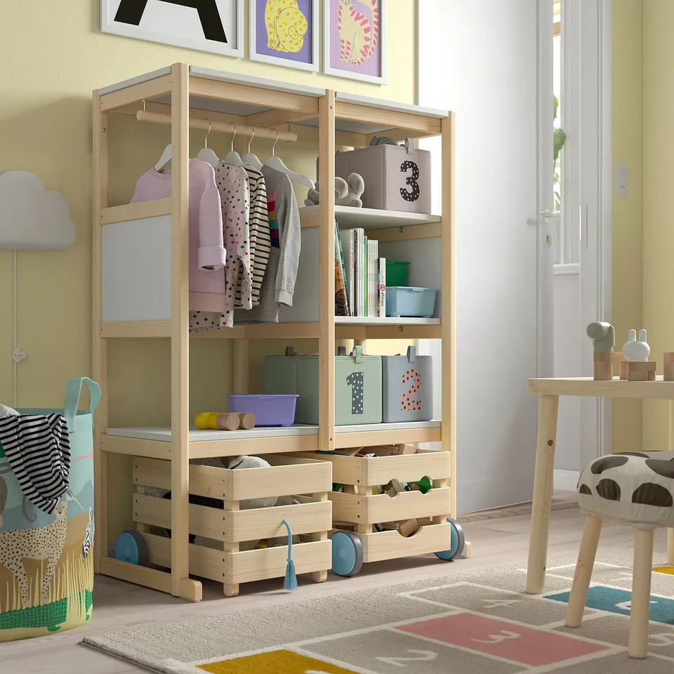 FLISAT light wood kids storage unit, with wheels and shelves, holding colourful boxes and hanging clothes, near a small table and a rug.