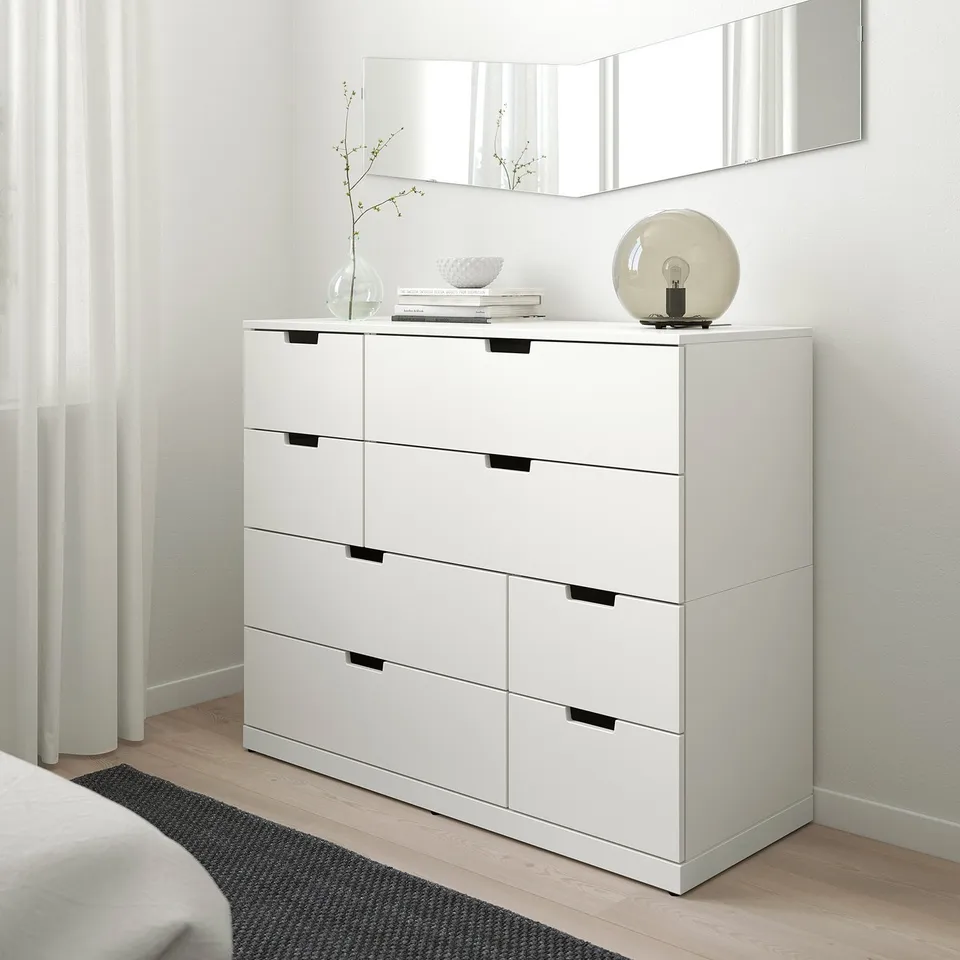 NORDLI chest with 6 drawers in modern bedroom. Beside bed, with lamp & vases. Sleek design, large mirror above, light floors & sheer curtains.