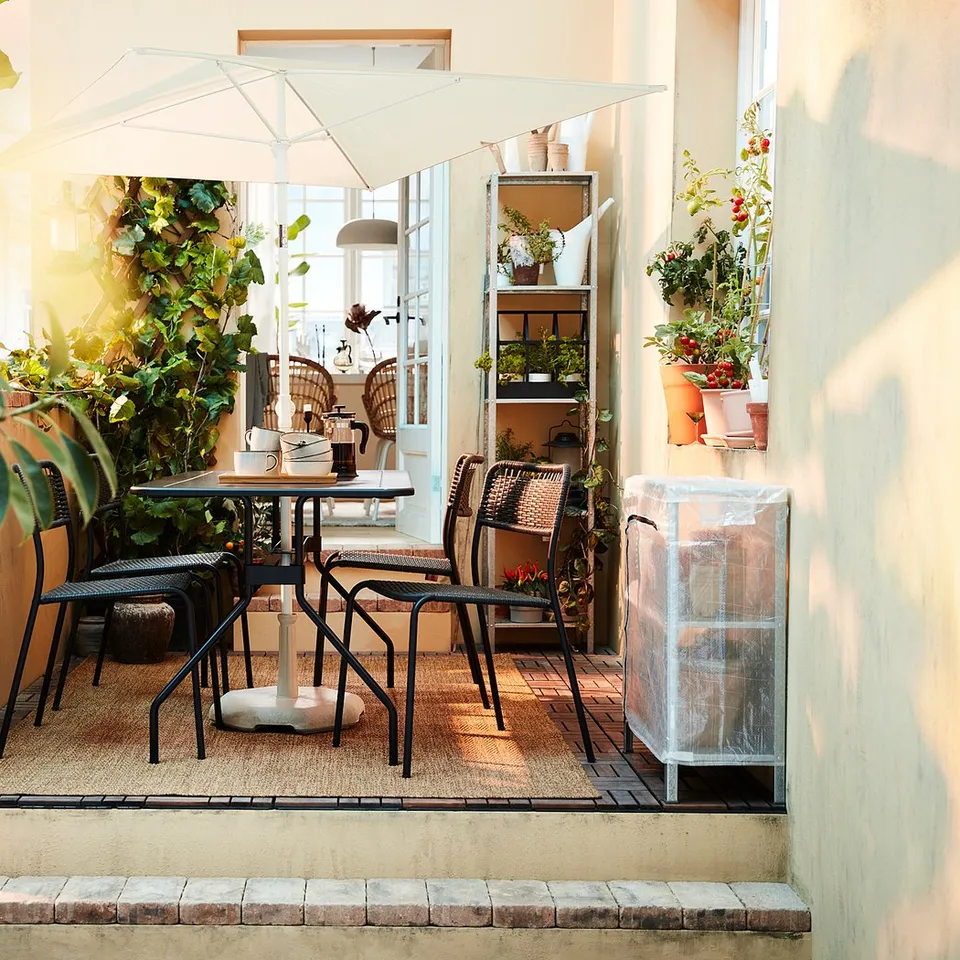 Small patio with black metal table and chairs, surrounded by potted plants. White adjustable TVETÖ parasol provides shade. Bright, cosy atmosphere.
