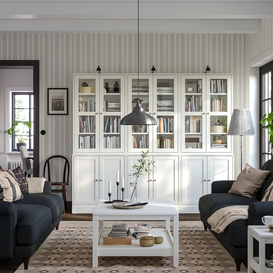 Modern living room with white HAVSTA shelving units filled with books and decor, grey couch, coffee table, patterned rug, and large windows.