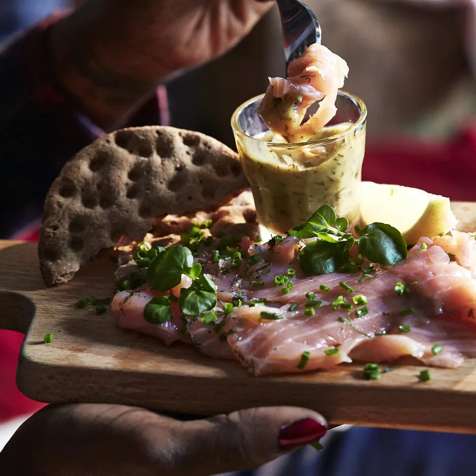 Hand holding smoked fish, greens, bread, and dipping sauce.
