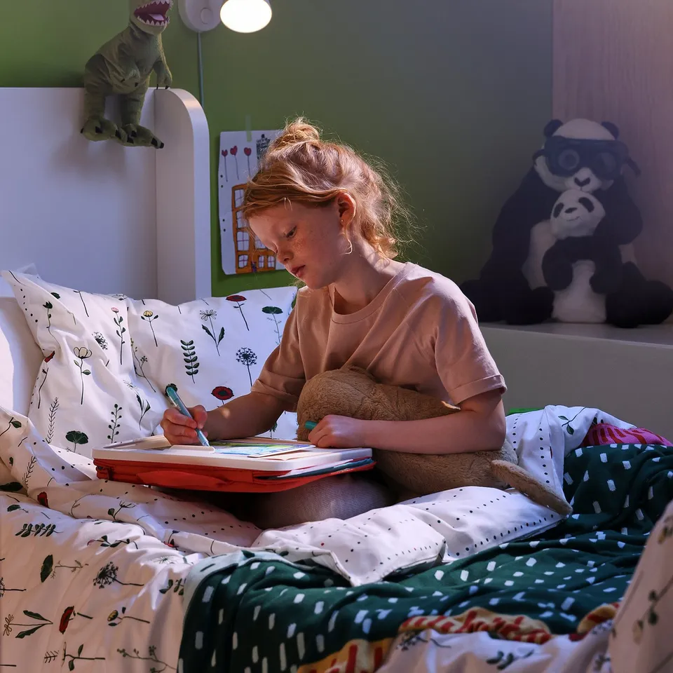 A girl sits on a bed with a green and floral NATTSLÄNDA duvet cover, drawing in a book. A dinosaur lamp is on, and a panda teddy bear is nearby.