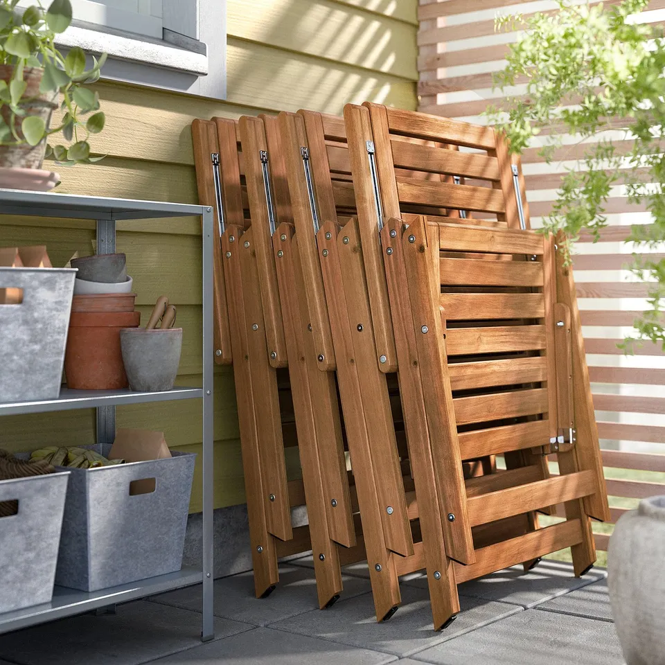 Brown wooden NÄMMARÖ folding chairs stacked outdoors next to storage shelves with potted plants.