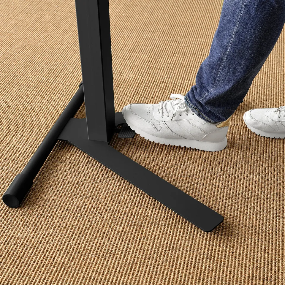 Person standing on a stylish black adjustable laptop table with white trainers and blue jeans on a patterned carpet.