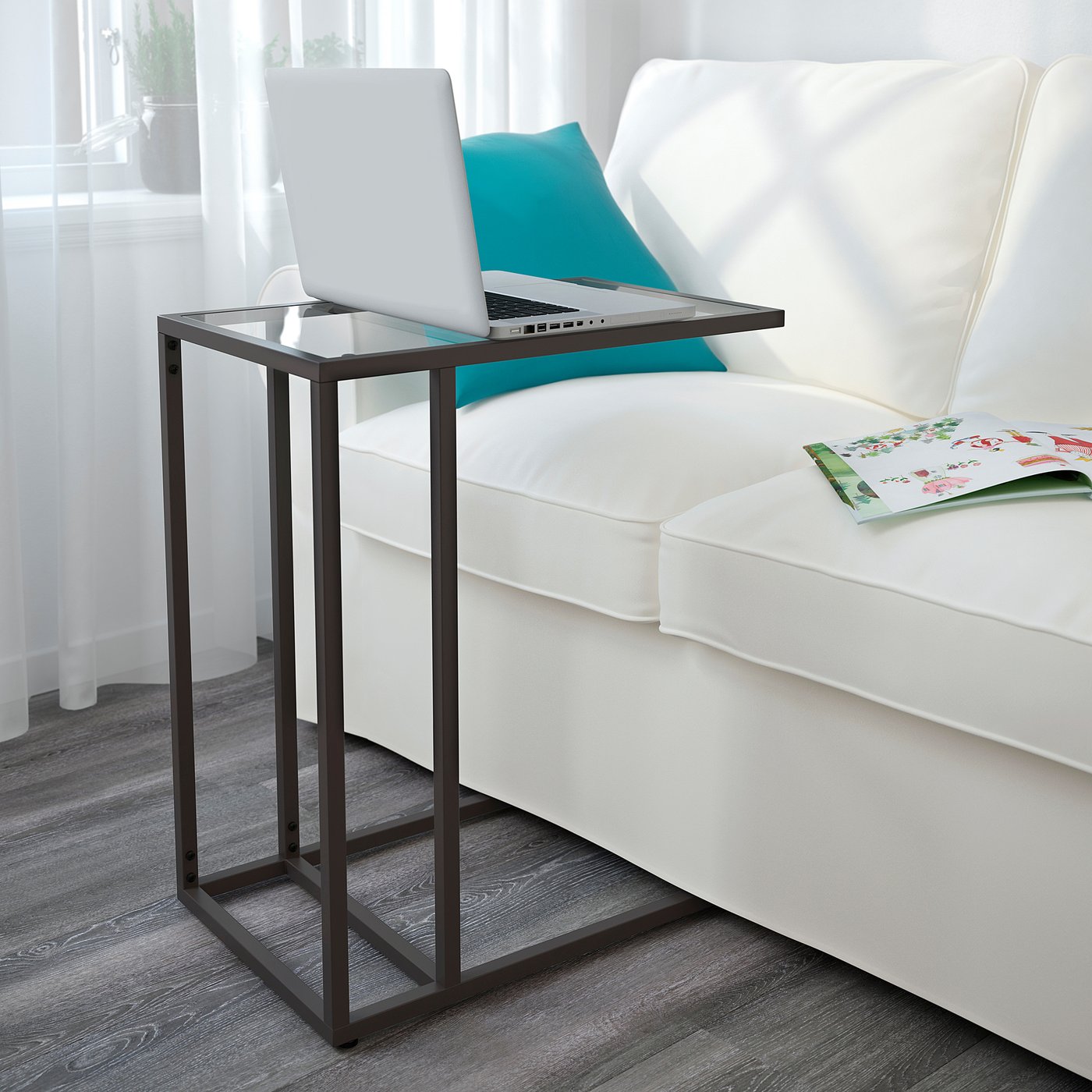 Small glass-top side table next to white couch, holding a laptop and a book.