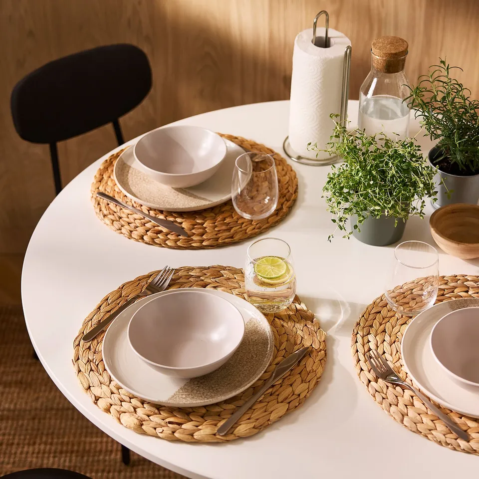 Three place settings with natural woven placemats, white bowls, plates, glasses, cutlery, and a centrepiece with fresh herbs on a white table.