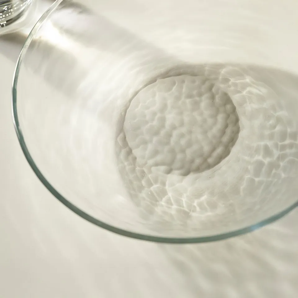Clear glass bowl with hammered texture, casting intricate shadow patterns, sits on a white surface.