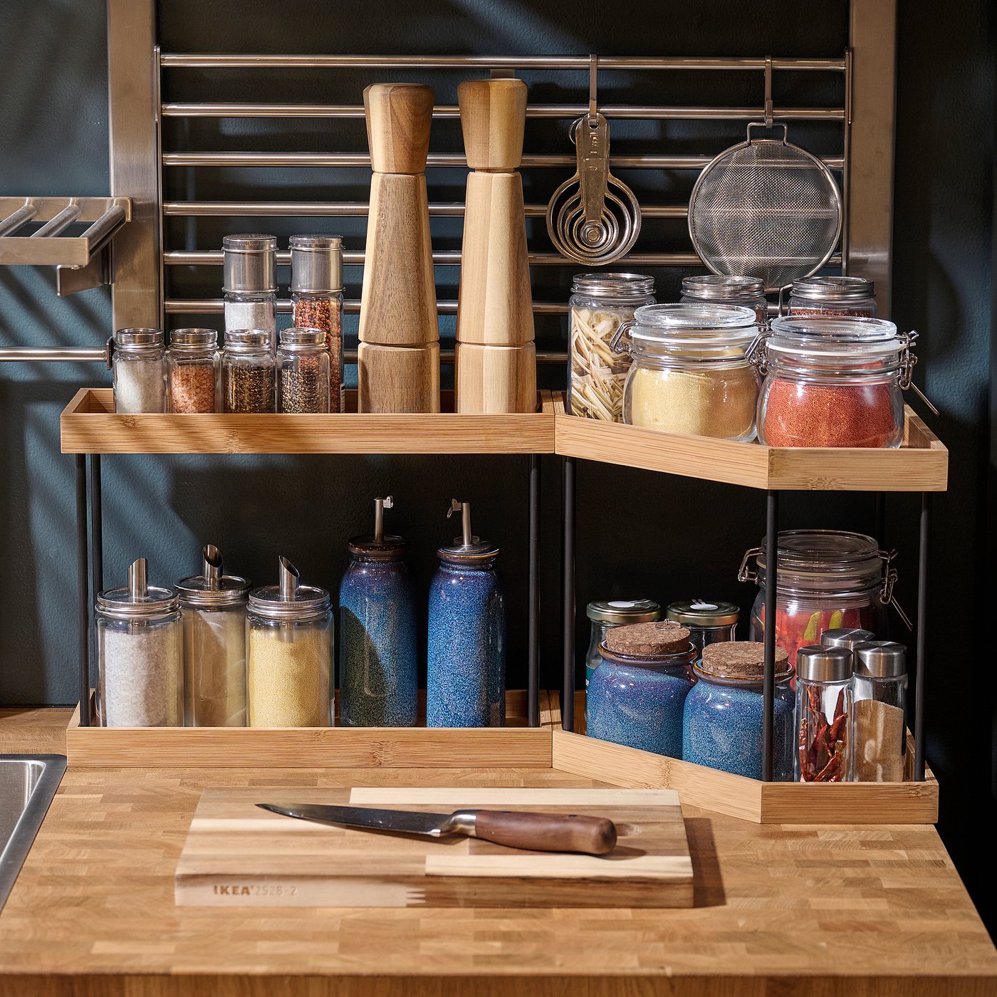 Well-organized kitchen storage. Bamboo shelves hold spices in jars, bottles, and mills on a countertop. Knife on cutting board is visible.