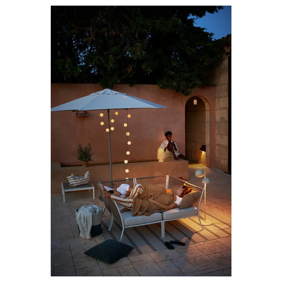 Patio with beige sofa, armchair, blue umbrella & lights. Person reading.