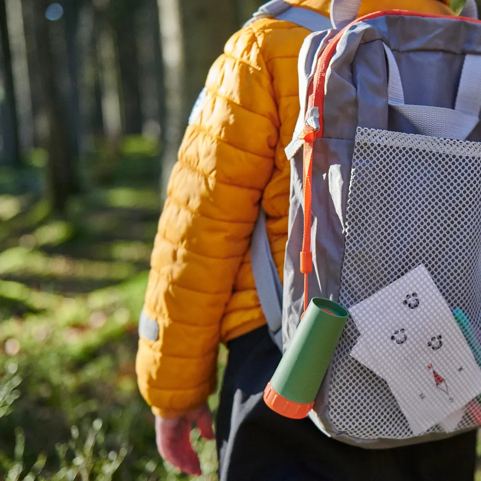 Child wearing bright orange jacket with blue MAJBAGGE backpack, carrying a green container and white card in mesh pocket.