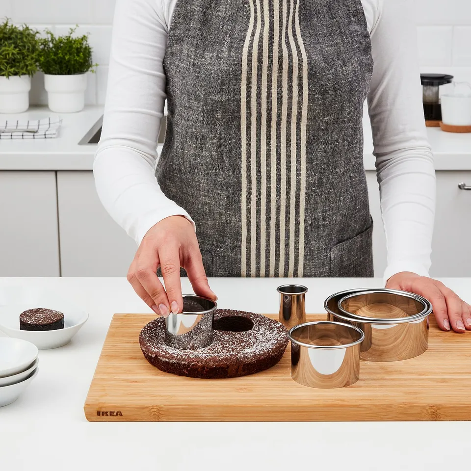 Person cuts cake with round cookie cutter on wooden board. Cylindrical cutters, plates, and a small cake also present.