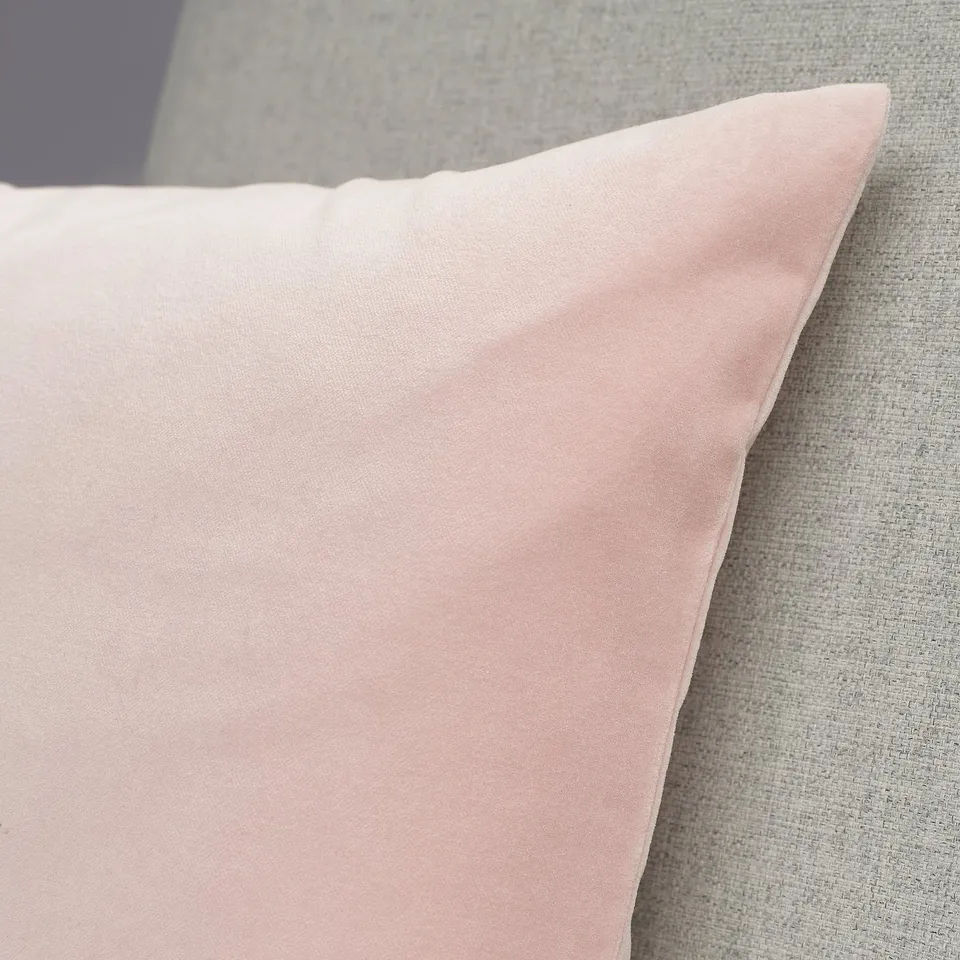 A close-up of a soft, light pink velvet cushion placed on a grey fabric surface.