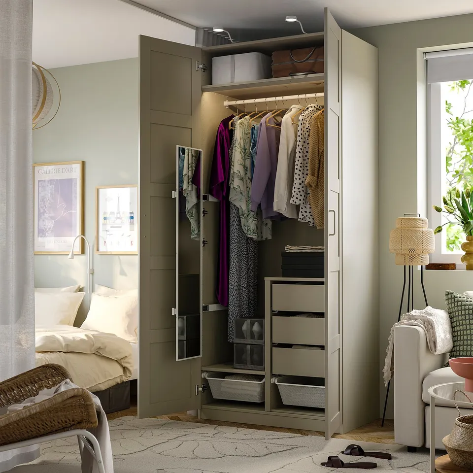 A bedroom with an open pax wardrobe displaying hanging clothes, pull-out shelves, and a built-in full-length mirror reflecting the room.