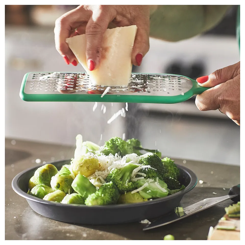 Person grates cheese with green UPPFYLLD grater onto bowl of broccoli and brussels sprouts.