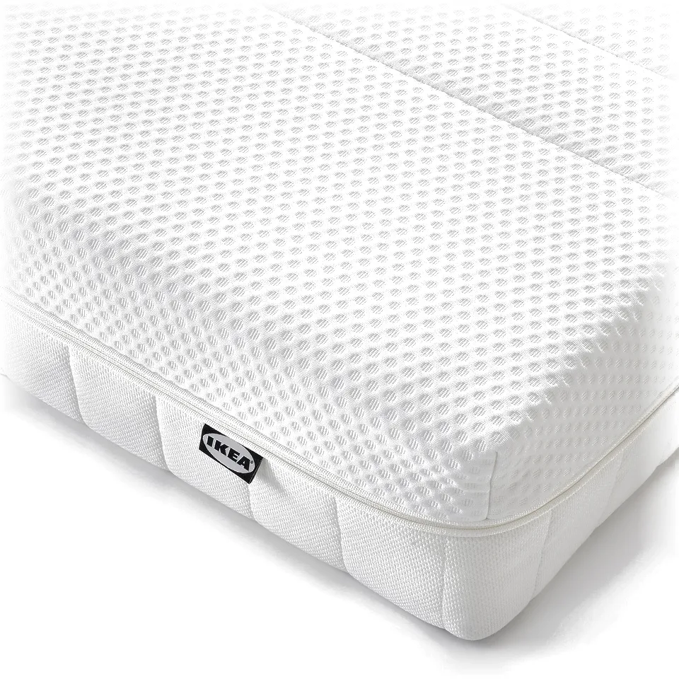 Corner of a white foam mattress with ikea label, featuring layered comfort zones and visible handles for moving.