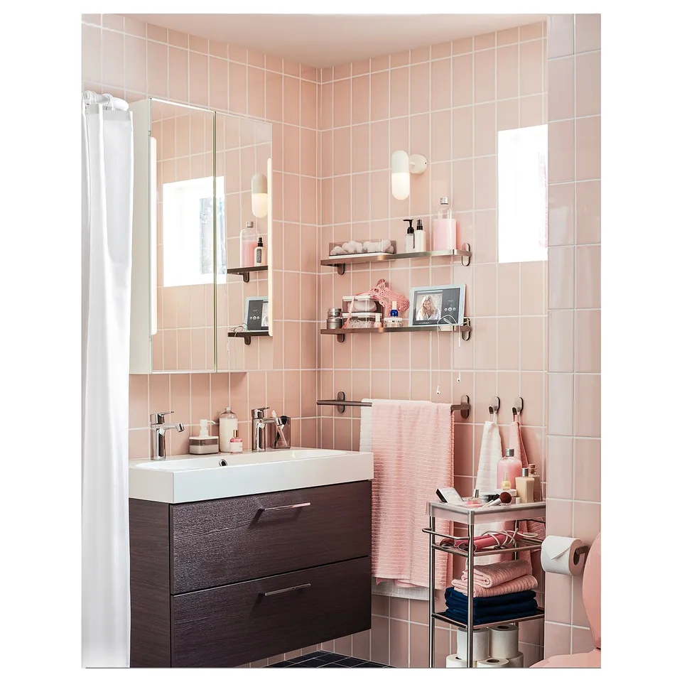 Modern bathroom with pink tiles, dark wood cabinet, white sink, wall shelves with bottles, towels, and decor, mirror with lights.