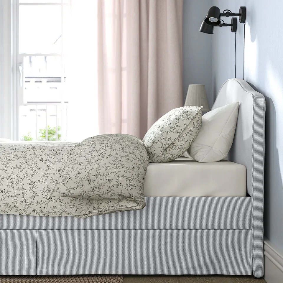 A cosy bedroom with a RAMNEFJÄLL bed, featuring a soft, curved headboard and striped bedding. A lamp is mounted on the wall beside the bed.