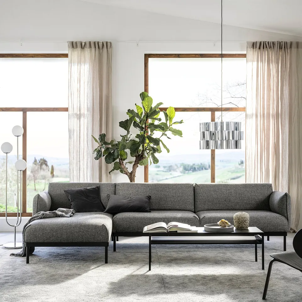 Minimalist living room with grey sofa, black cushions, wooden table, lamp, and window view.