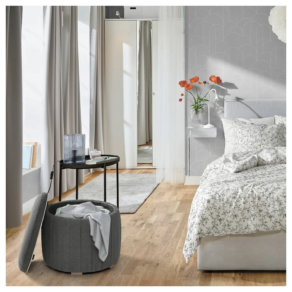 Modern bedroom with ÅTERLÖGA grey fabric pouffe for storage, round side table, wooden floor, floral bedding, and large windows with draped curtains.