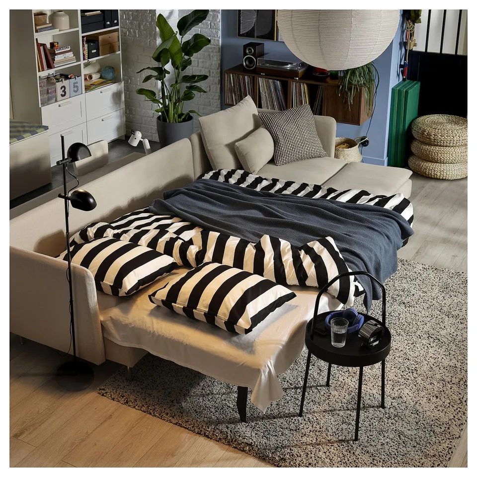 Modern living room with beige sofa bed, striped pillows, coffee table, rug, plants, and bookshelf.