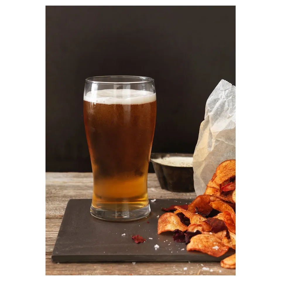 Clear glass beer glass with foam, sitting on slab next to crispy snacks and salt shaker.
