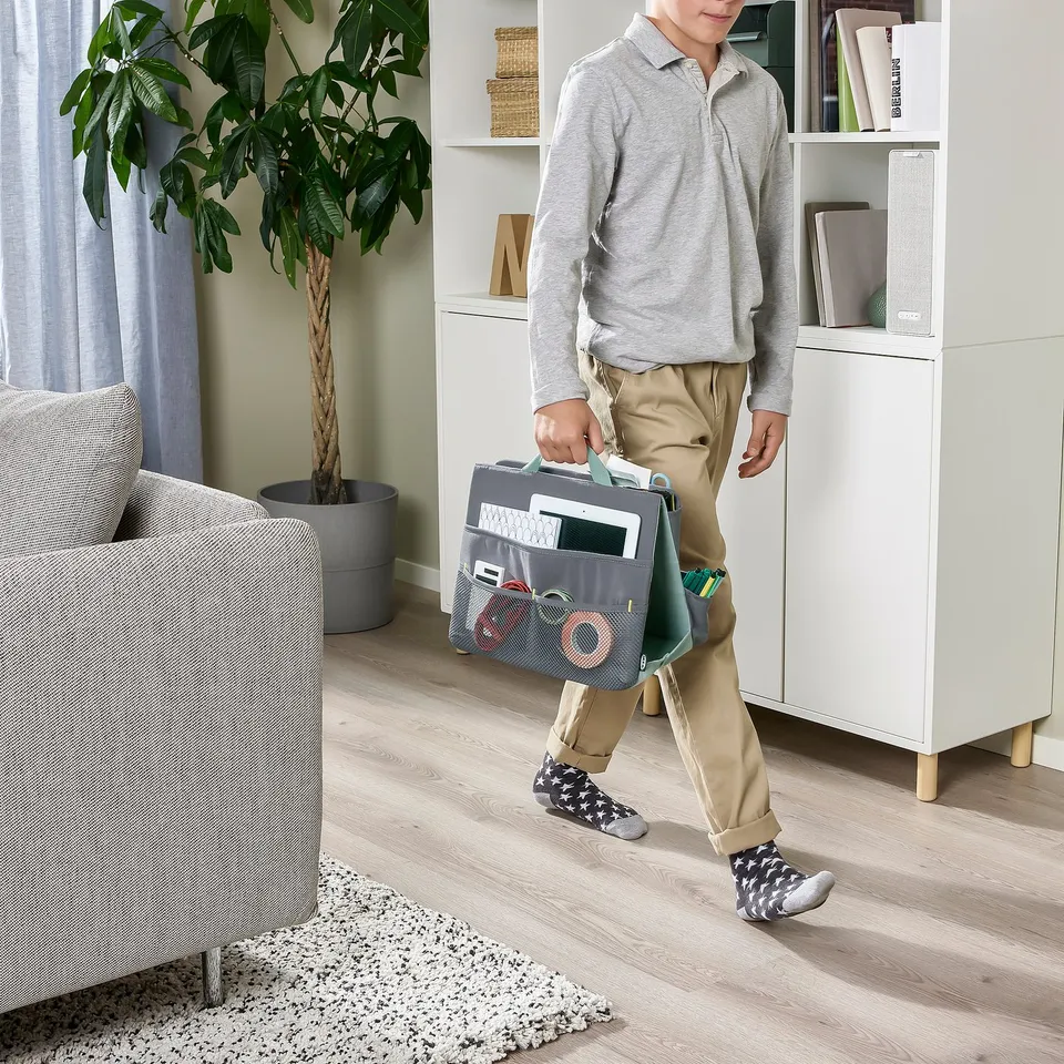 Person holding an ÖVNING organiser, which carries school supplies and a tablet, walking in a room with a sofa and shelves.