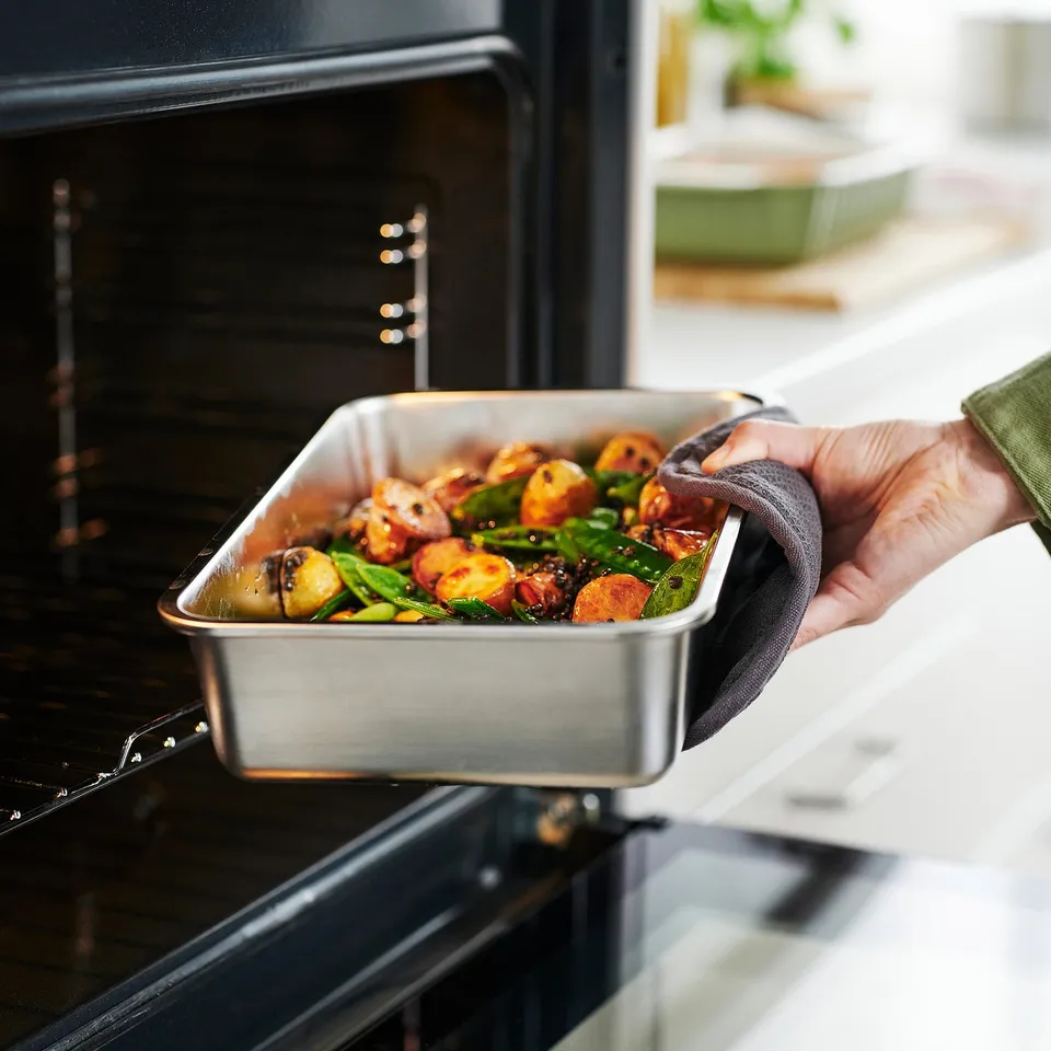 Hand holding metal tray with roasted vegetables, being placed into an open black oven.