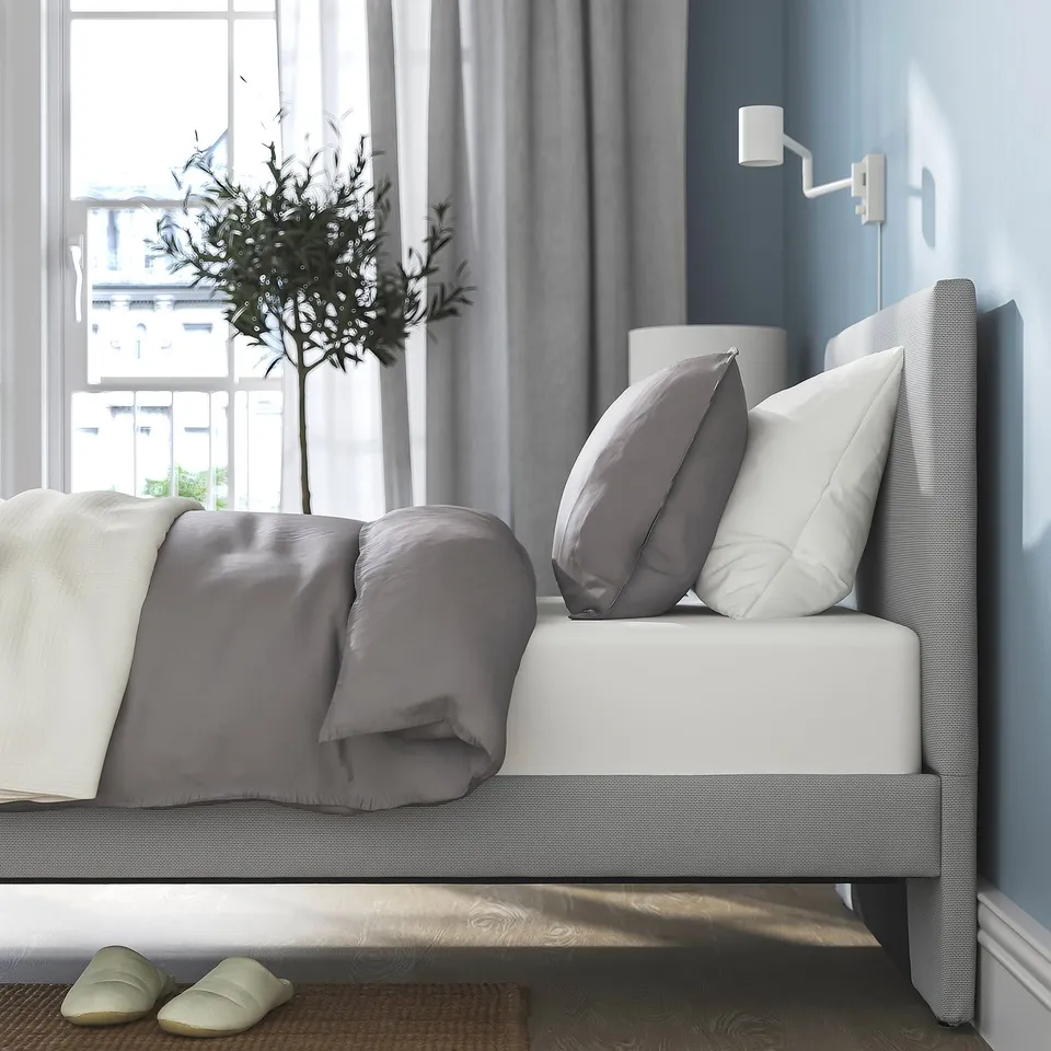 Cosy bedroom with grey GLADSTAD bed, soft headboard, white pillows, grey blanket, slippers, plant, curtain, wall lamp.