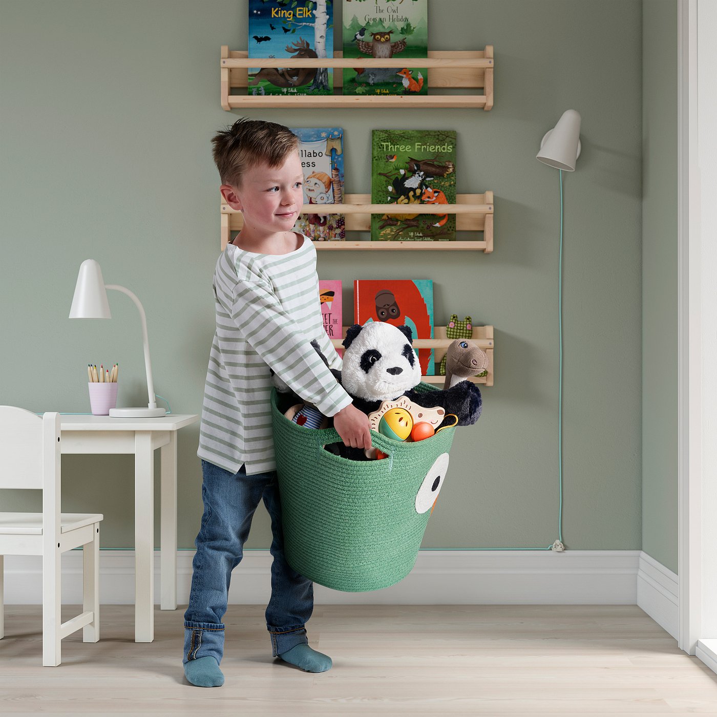 Boy holding green storage basket with stuffed panda, standing near shelves with books and a desk.