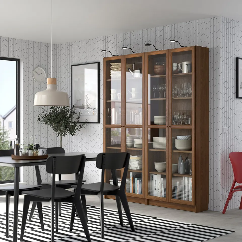 Modern dining room with a tall wooden OXBERG bookcase, glass doors, dishes, and a black dining set. Large plant and striped rug.