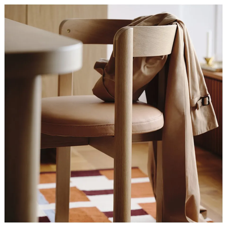 Modern dining chair in oak wood with upholstered seat, tan leather jacket draped over back.