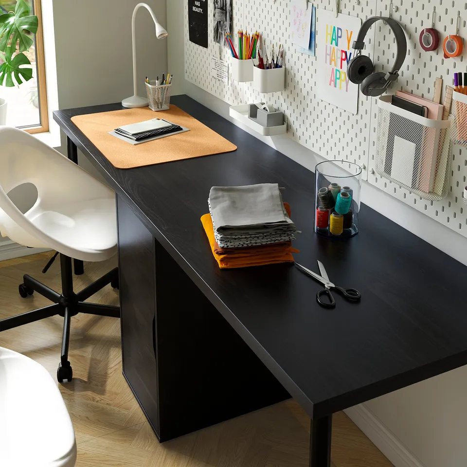 Black desk with chair, lamp, laptop, stationery, fabric, scissors, and pens organised on pegboard.
