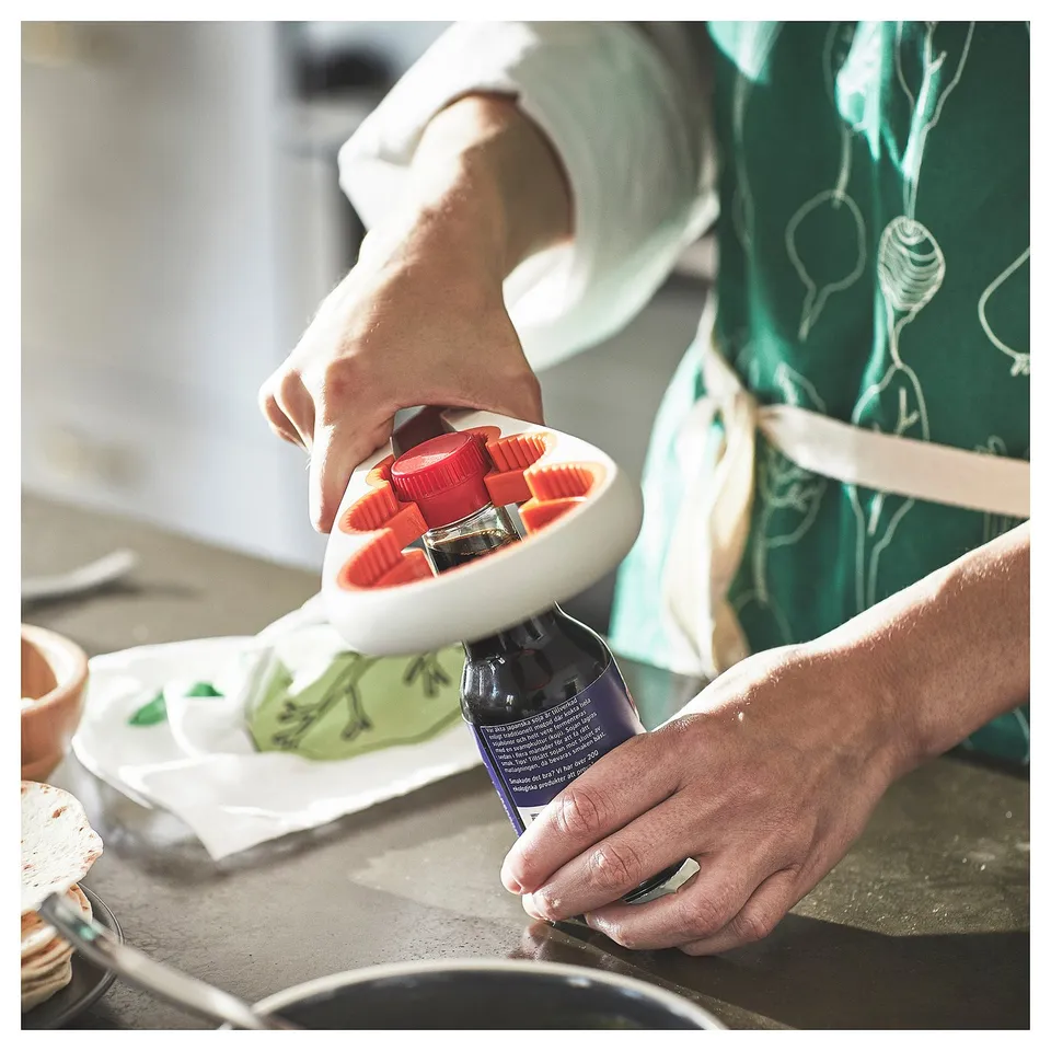 Person using red UPPFYLLD jar opener on glass jar. Green and white apron, kitchen setting.