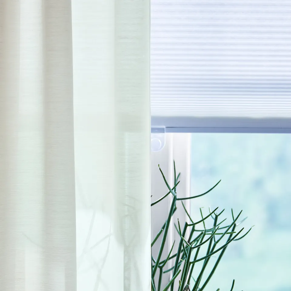 HOPPVALS white blind partially covers window, green plant touches it, sunlight philtres through.