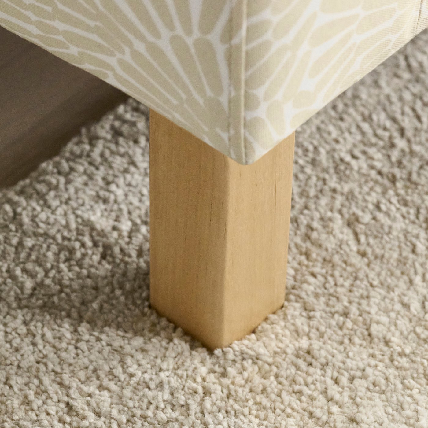 A close-up of a sofas corner with light fabric featuring a white pattern and a light wooden leg on a textured carpet.