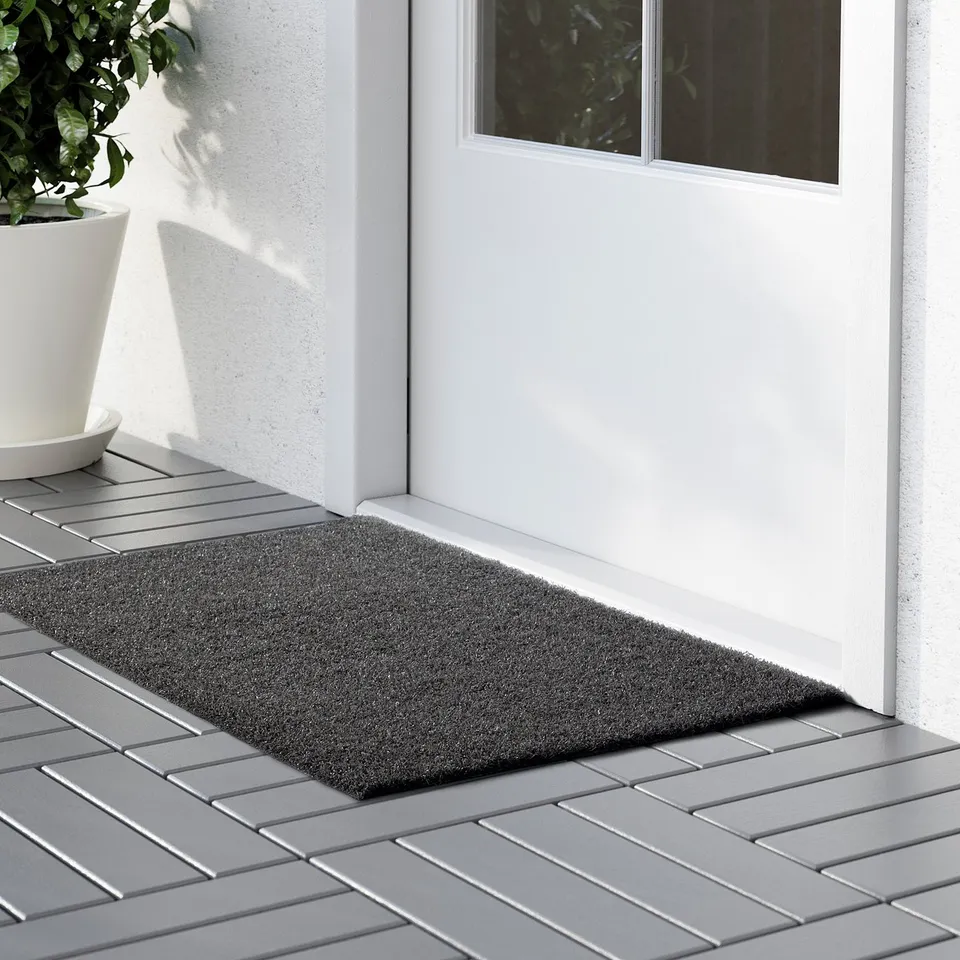 A rectangular doormat placed outside a white door, designed to withstand weather and dirt, with a non-slip backing visible beneath it.