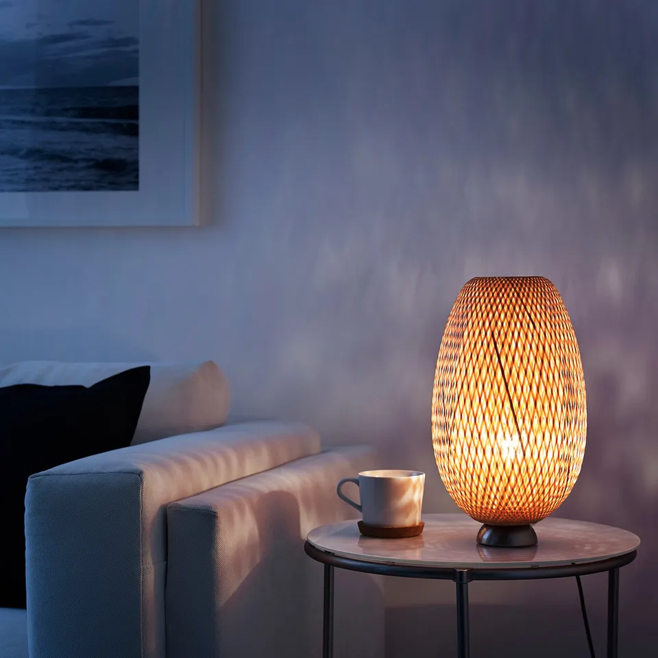 A cosy living room with a lit table lamp, casting warm light and patterns on the wall. Next to a couch with pillows and a mug on a side table.