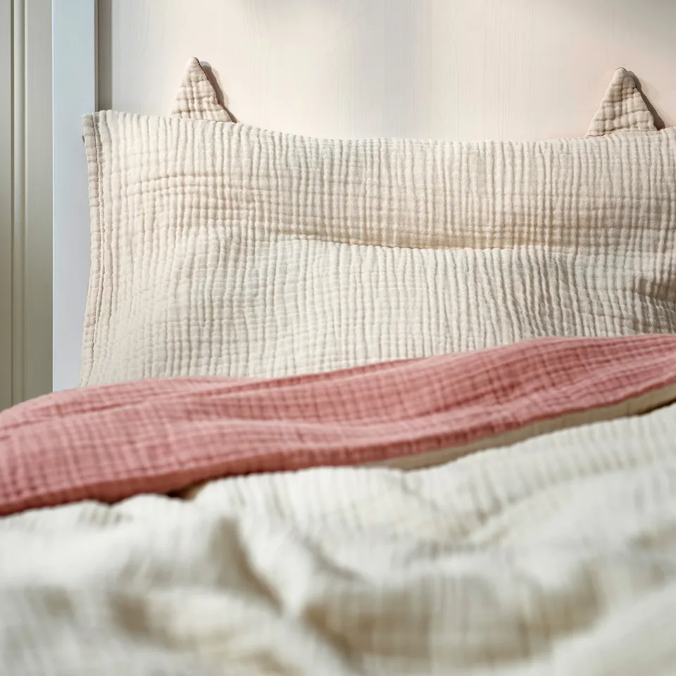 Soft, baby bedding set in light pink with white trim, featuring a pillow with ears. Made of organic cotton.