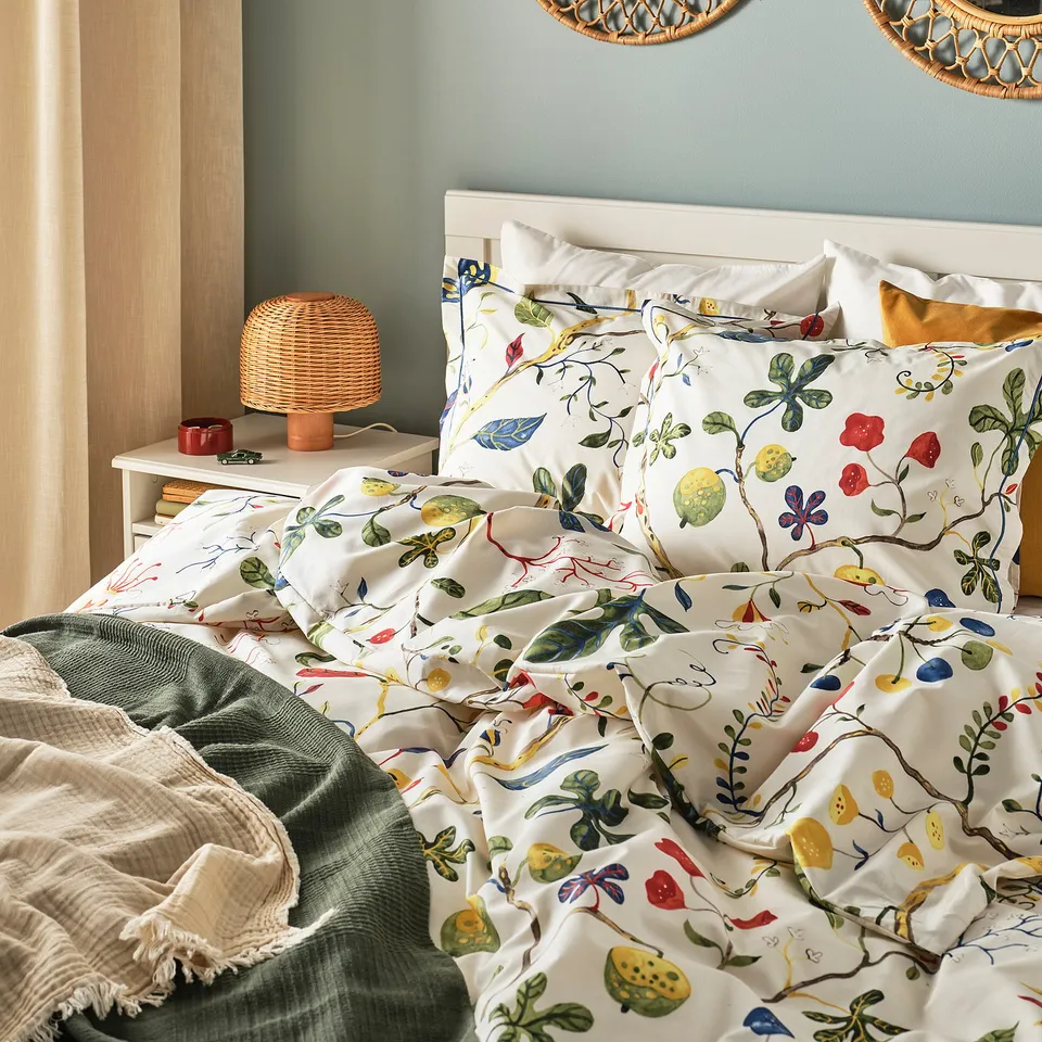 Cosy bed with KRONBUSKE floral duvet, wooden headboard, wicker lamp, and green blanket.